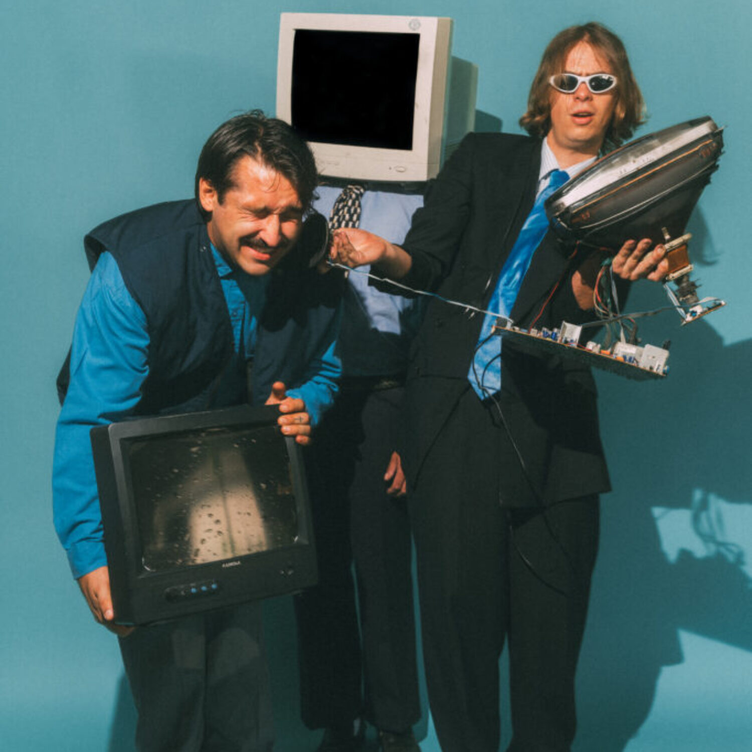 083_Bild_10-Musik_Tiavo_FR-20-30_mxm-photo Die drei Bandmitglieder der Band Tiavo sind mitten in der Bewegung vor einem blauen Hintergrund abgelichtet. Sie tragen Kleidung im Stil der 90er Jahre. Die beiden vorderen Bandmitglieder halten die Komponenten eines alten Röhrenfernsehers in der Hand, während die hintere Person einen Röhrenfernseher über den Kopf gestülpt hat.
